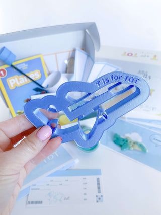 Complete airplane activity kit for kids featuring playdough cutter, airplane construction sets, art supplies for Into the Sky craft, airplane launch pad, steam and recipe cards, and measuring tape for educational activities.